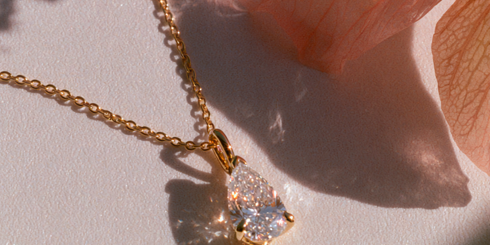 A gold chain necklace with a pear-cut clear gemstone pendant, styled on a white surface with soft peach flower petals and natural shadows.