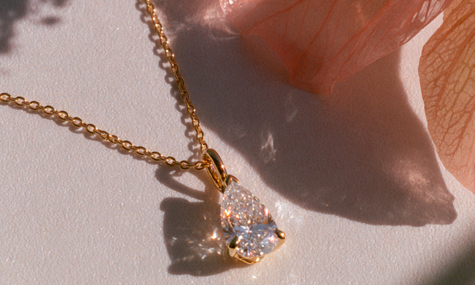 A gold chain necklace with a pear-cut clear gemstone pendant, styled on a white surface with soft peach flower petals and natural shadows.