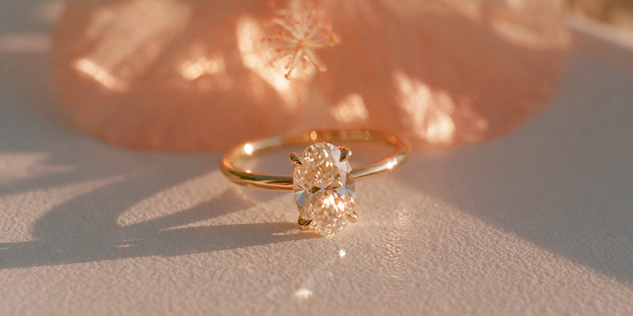 A gold ring with a large clear gemstone in a prong setting, resting on a textured light surface with a soft peach flower blurred in the background.