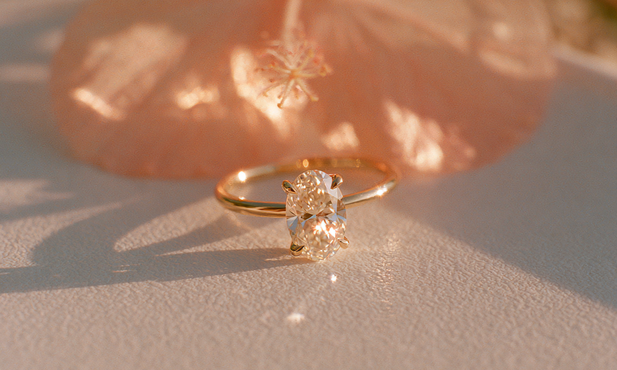 A gold ring with a large clear gemstone in a prong setting, resting on a textured light surface with a soft peach flower blurred in the background.