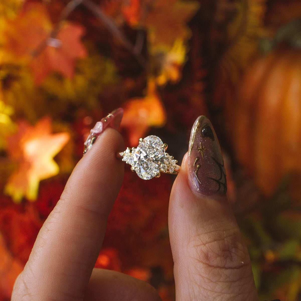 A close-up of a hand with glittery manicured nails holding a nova oval engagement ring with a large clear center stone and smaller surrounding stones, set against a warm autumn background of orange leaves and pumpkins.