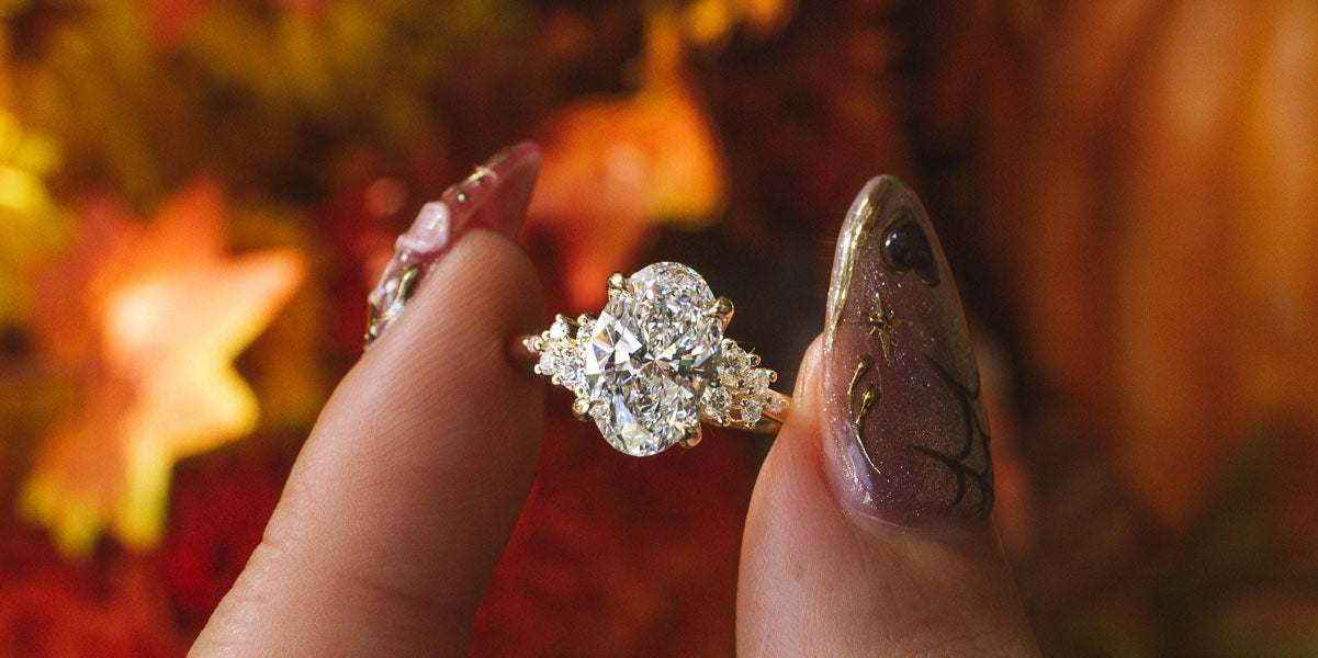 A close-up of a hand with glittery manicured nails holding a nova oval engagement ring with a large clear center stone and smaller surrounding stones, set against a warm autumn background of orange leaves and pumpkins.