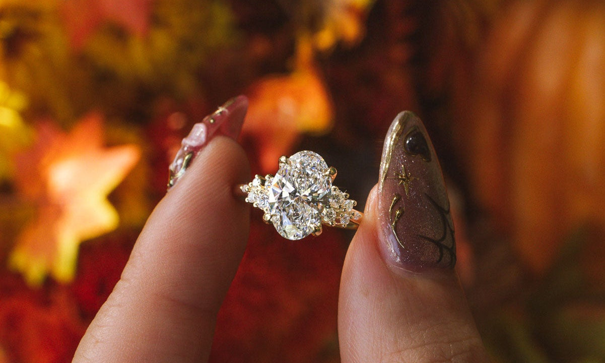 A close-up of a hand with glittery manicured nails holding a nova oval engagement ring with a large clear center stone and smaller surrounding stones, set against a warm autumn background of orange leaves and pumpkins.