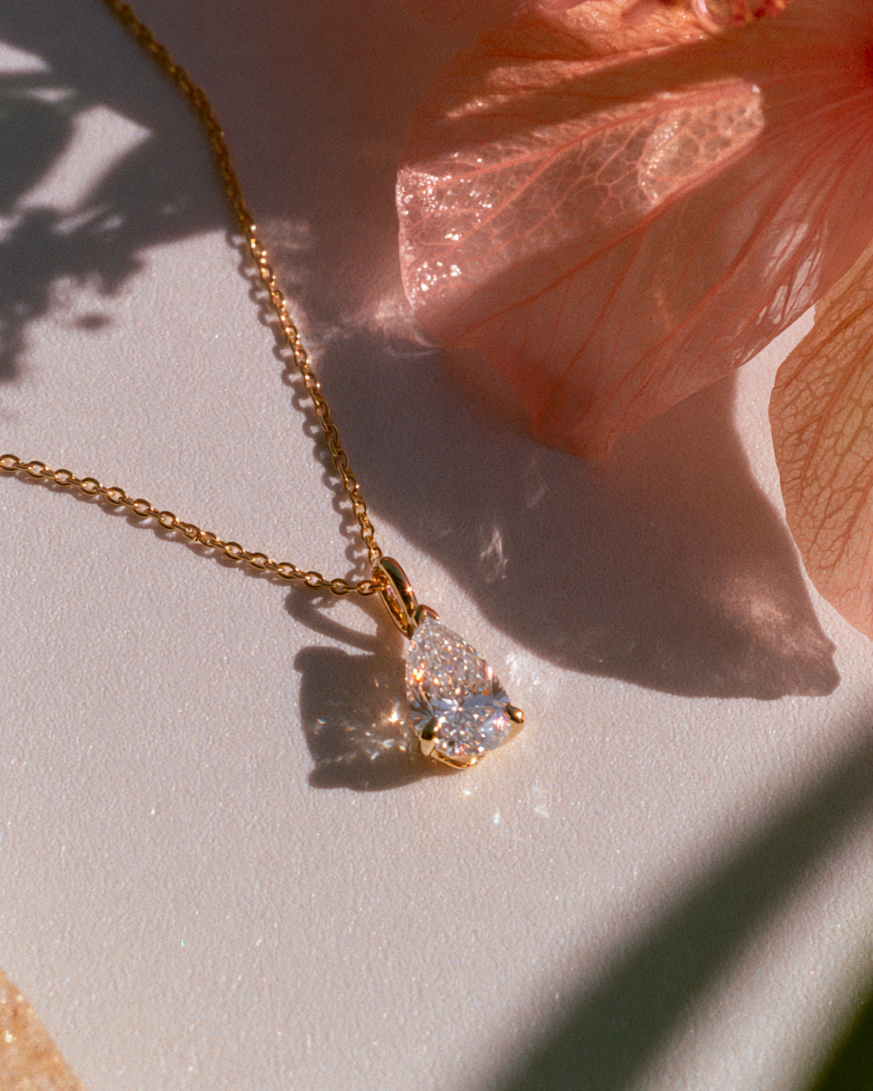 A gold chain necklace with a pear-cut clear gemstone pendant, styled on a white surface with soft peach flower petals and natural shadows.