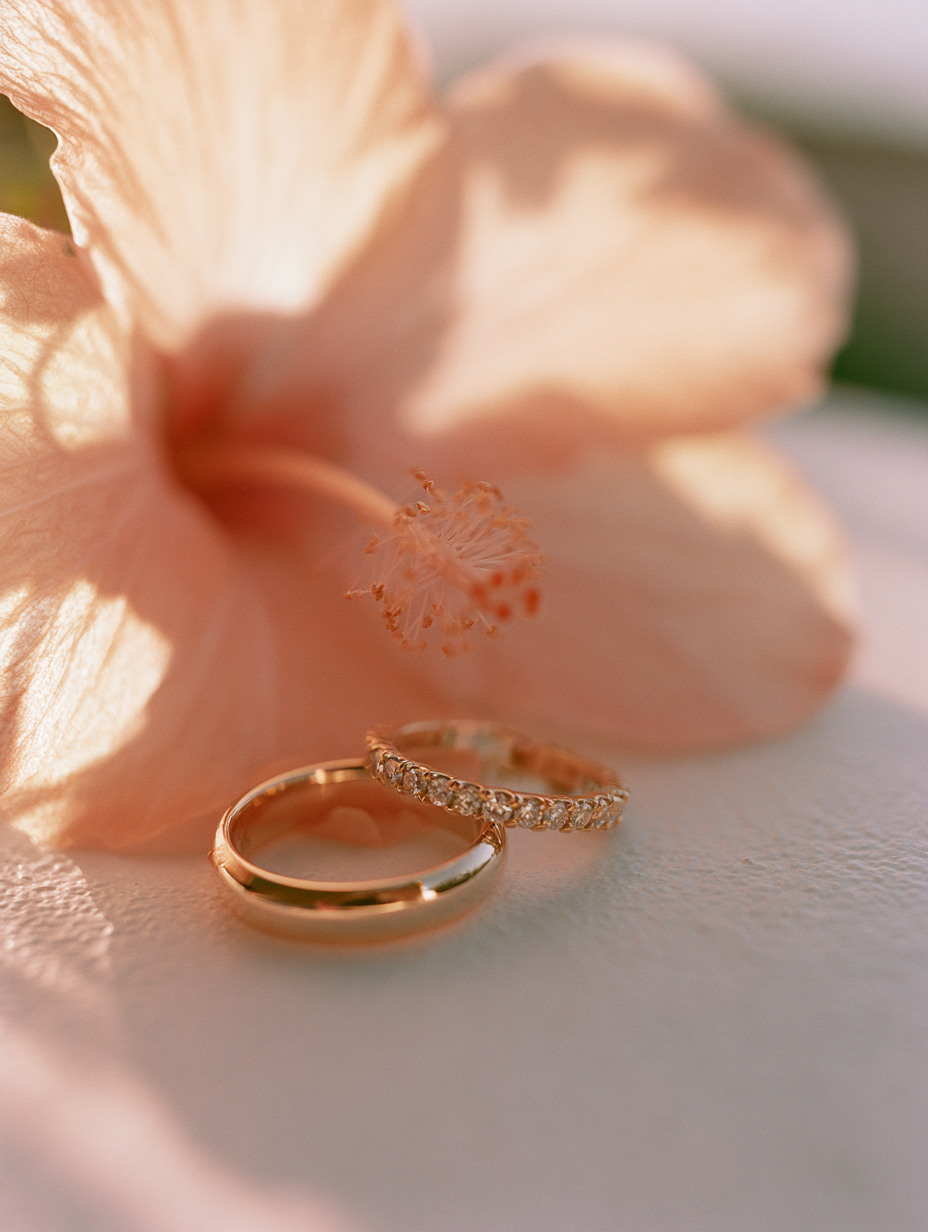 Two gold rings, one plain and one with a row of small clear stones, resting on a white surface in front of a soft peach hibiscus flower.