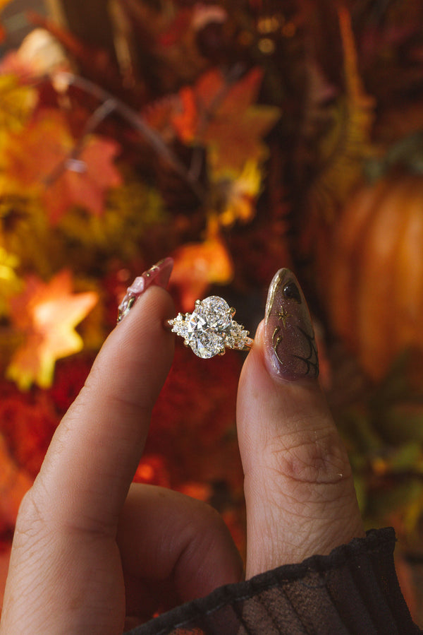 A close-up of a hand with glittery manicured nails holding a nova oval engagement ring with a large clear center stone and smaller surrounding stones, set against a warm autumn background of orange leaves and pumpkins.