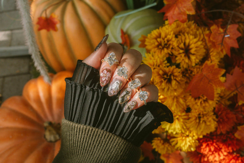 A close-up of a hand wearing a black pleated knit glove, showcasing long manicured nails decorated with glittery silver and white floral patterns. The background is a cozy autumn scene featuring pumpkins, orange mums, and fall leaves.